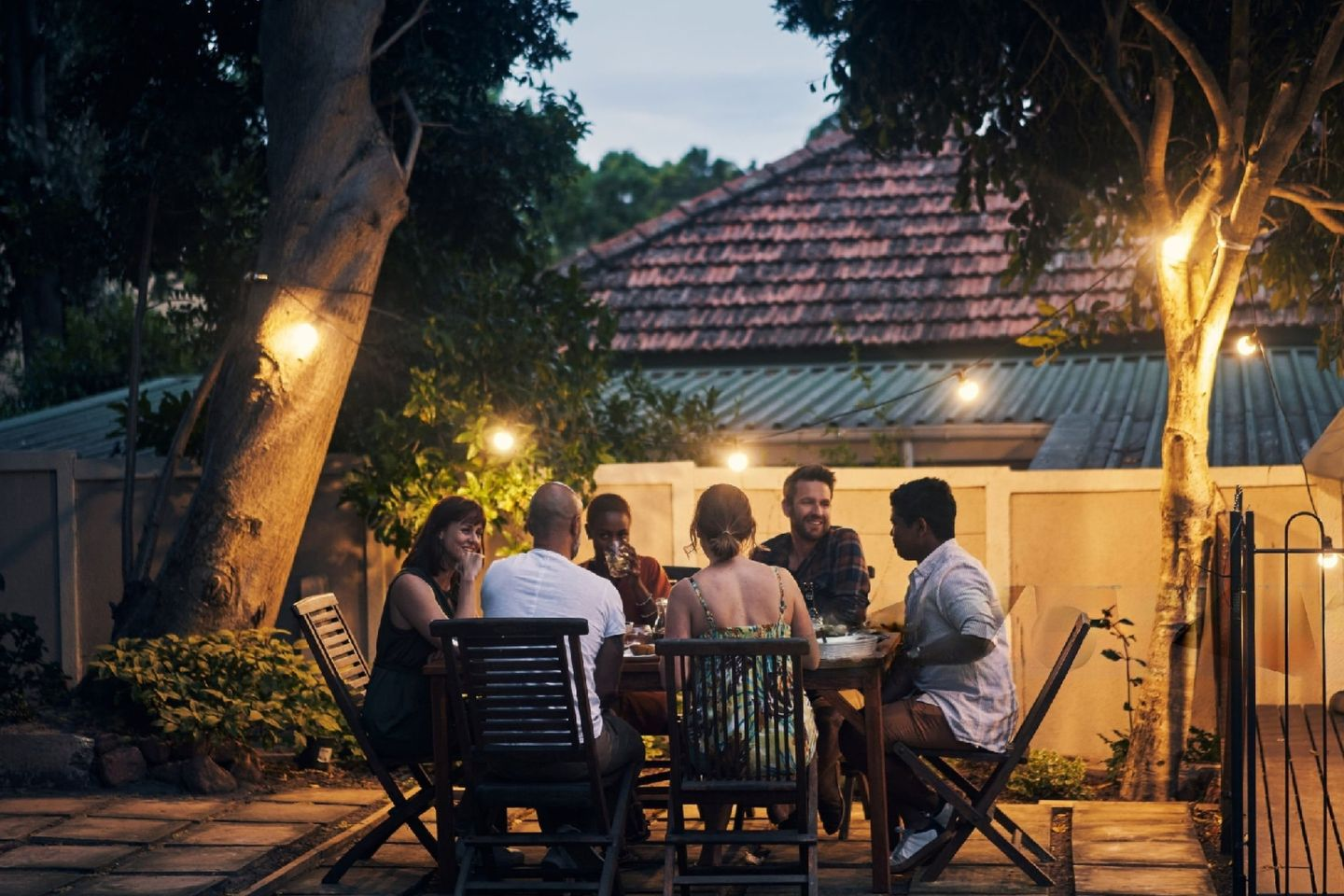 Groupe d'amis souriants et discutant, assis à table sur une terrasse de jardin en bois, sous la lumière d'une guirlande de style guinguette.