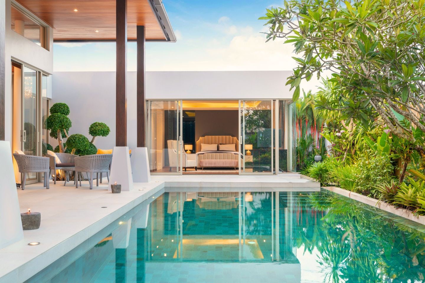 Une piscine à l'eau limpide de face, qui se prolonge sur une chambre parentale avec baie vitrée. Sur la gauche, terrasse avec table et chaises en rotin, à droite, végétation tropicale.